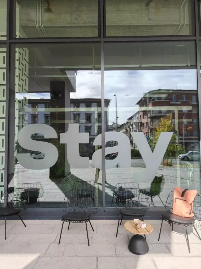 Vetrata d’ingresso del the social hub florence belfiore con sedute esterne e scritta “stay”.